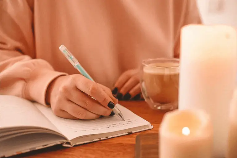 Mulher escrevendo em um caderno com uma xícara de café ao lado, em um ambiente acolhedor com velas acesas.