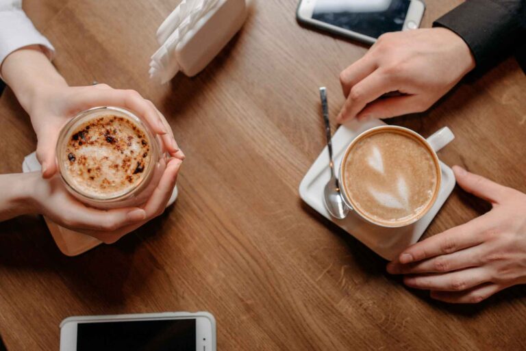 Duas xícaras de café sobre a mesa com relógio, vela e canela ao fundo, simbolizando conexão, amizade e o tempo de uma boa conversa.